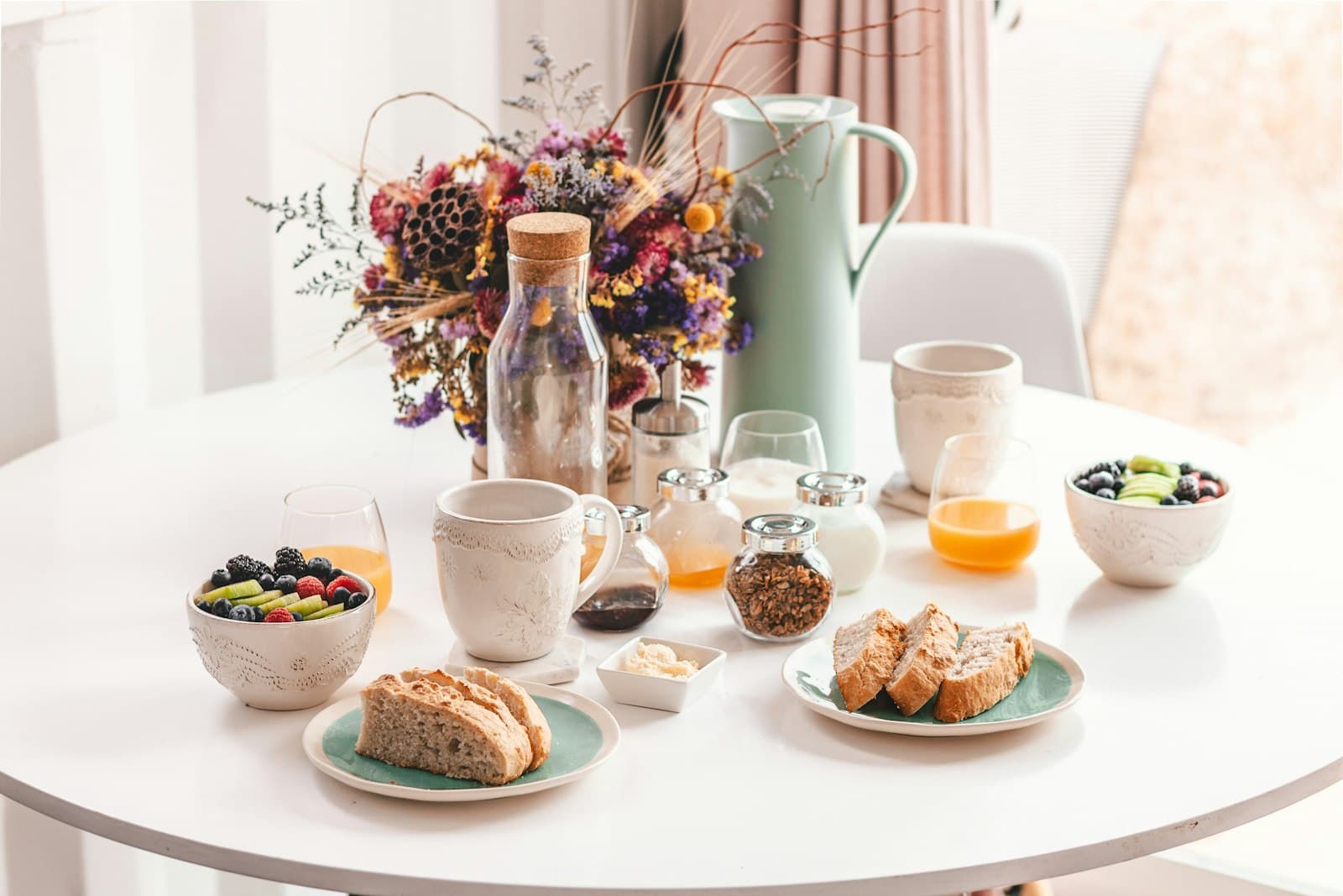 Desayuno en una mesa redonda blanca: pan, fruta, café y una jarra con flores secas