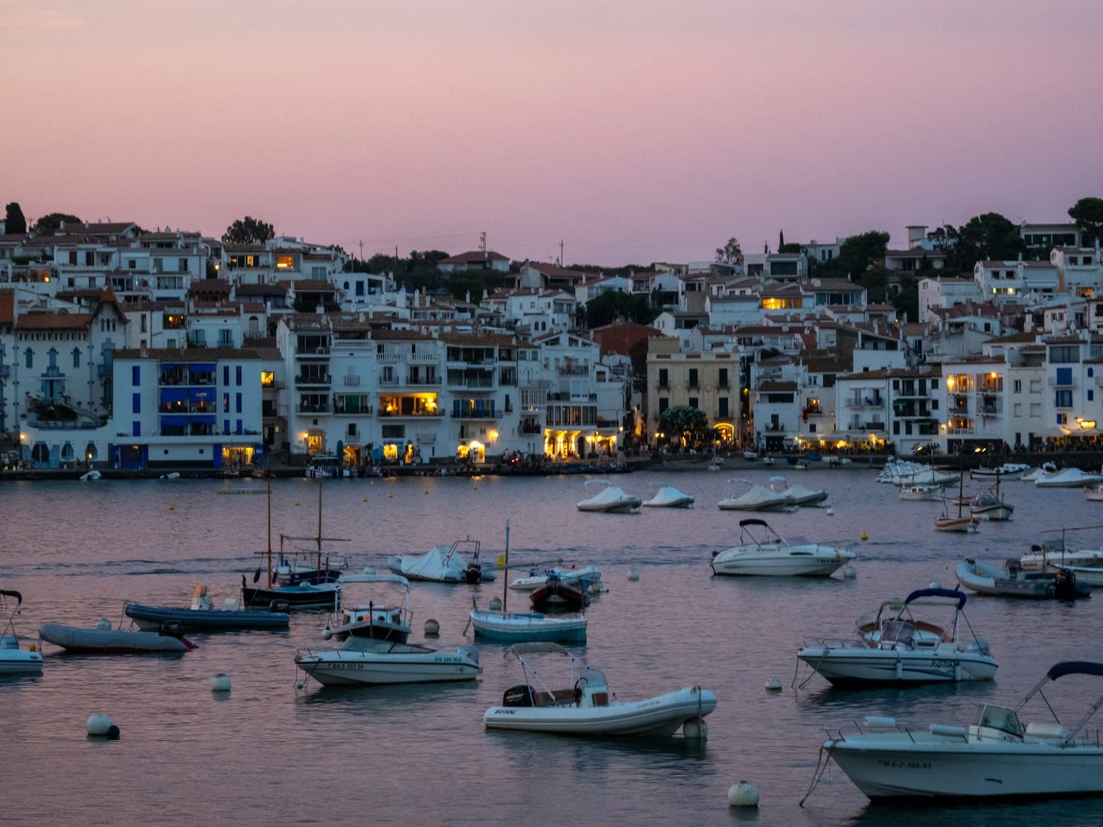 El puerto al anochecer: barcas pequeñas sobre un agua rosa y quieta, y las luces del pueblo blanco empezando a encenderse