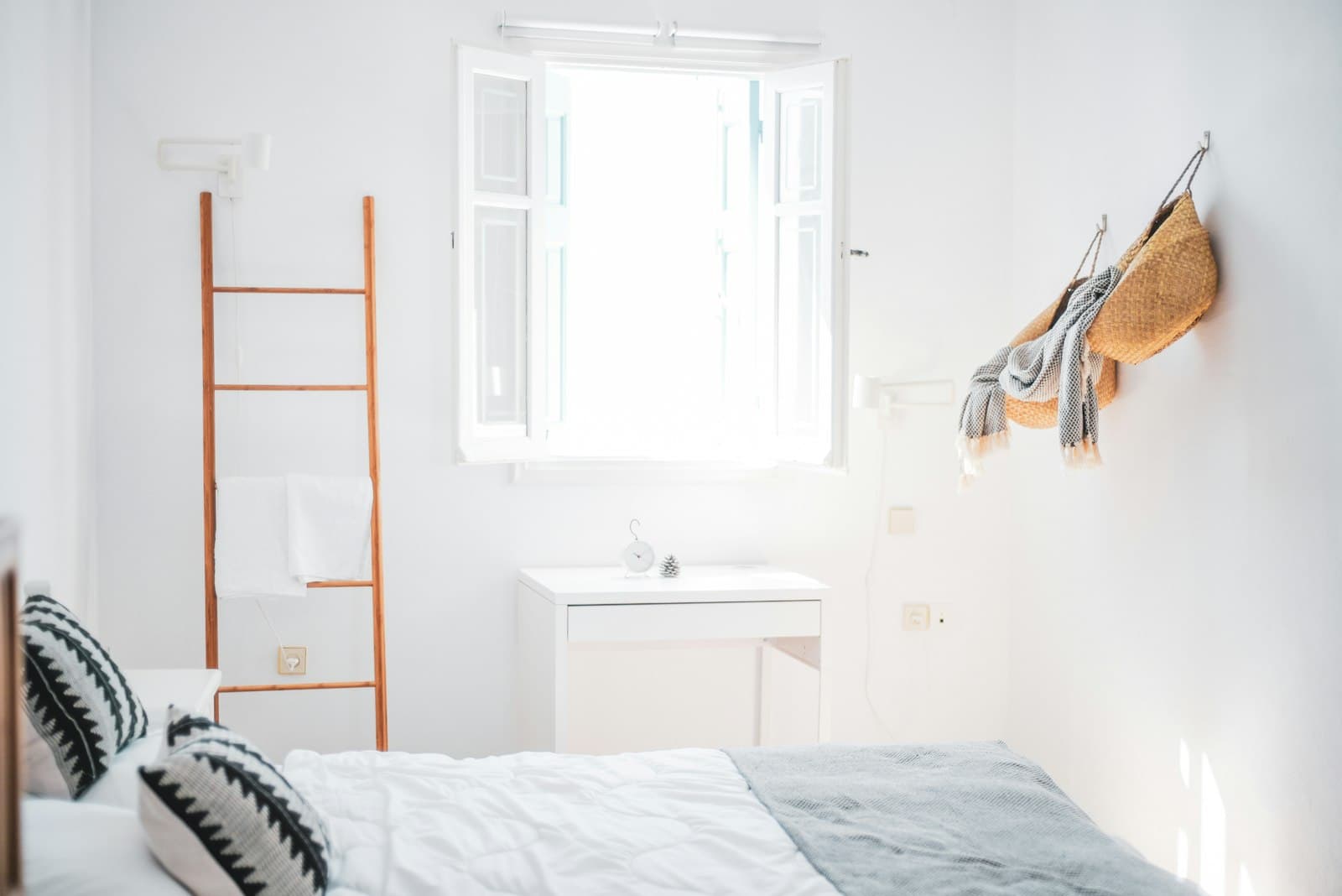 Habitación blanca con la contraventana abierta, cama vestida de lino y una escalera de madera para las toallas
