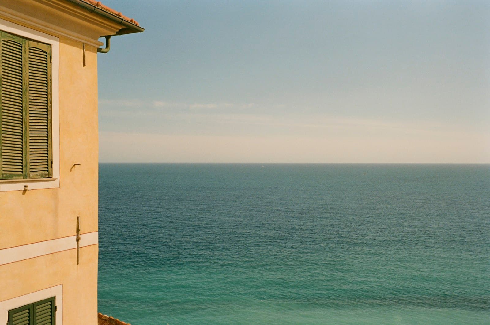 Un muro ocre con contraventanas verdes frente a un mar en calma y vacío