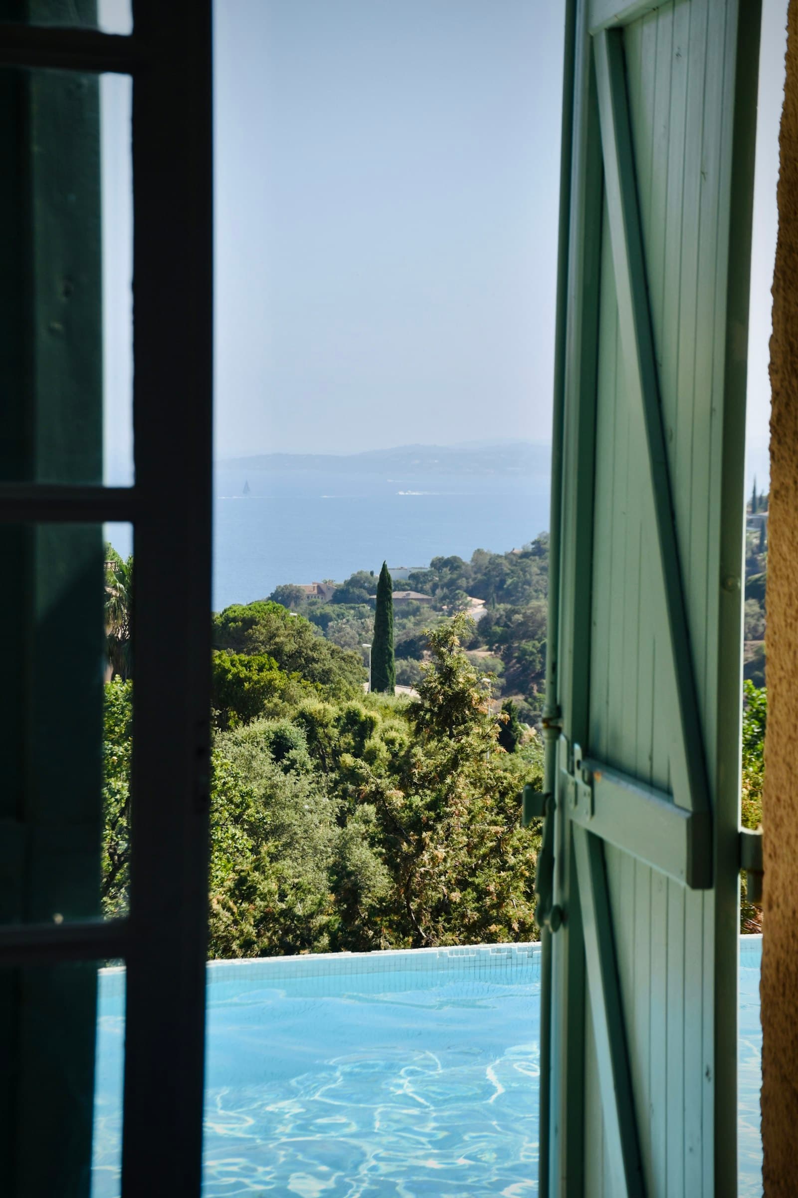 Contraventanas verdes abiertas a la piscina, con los pinos y el mar al fondo