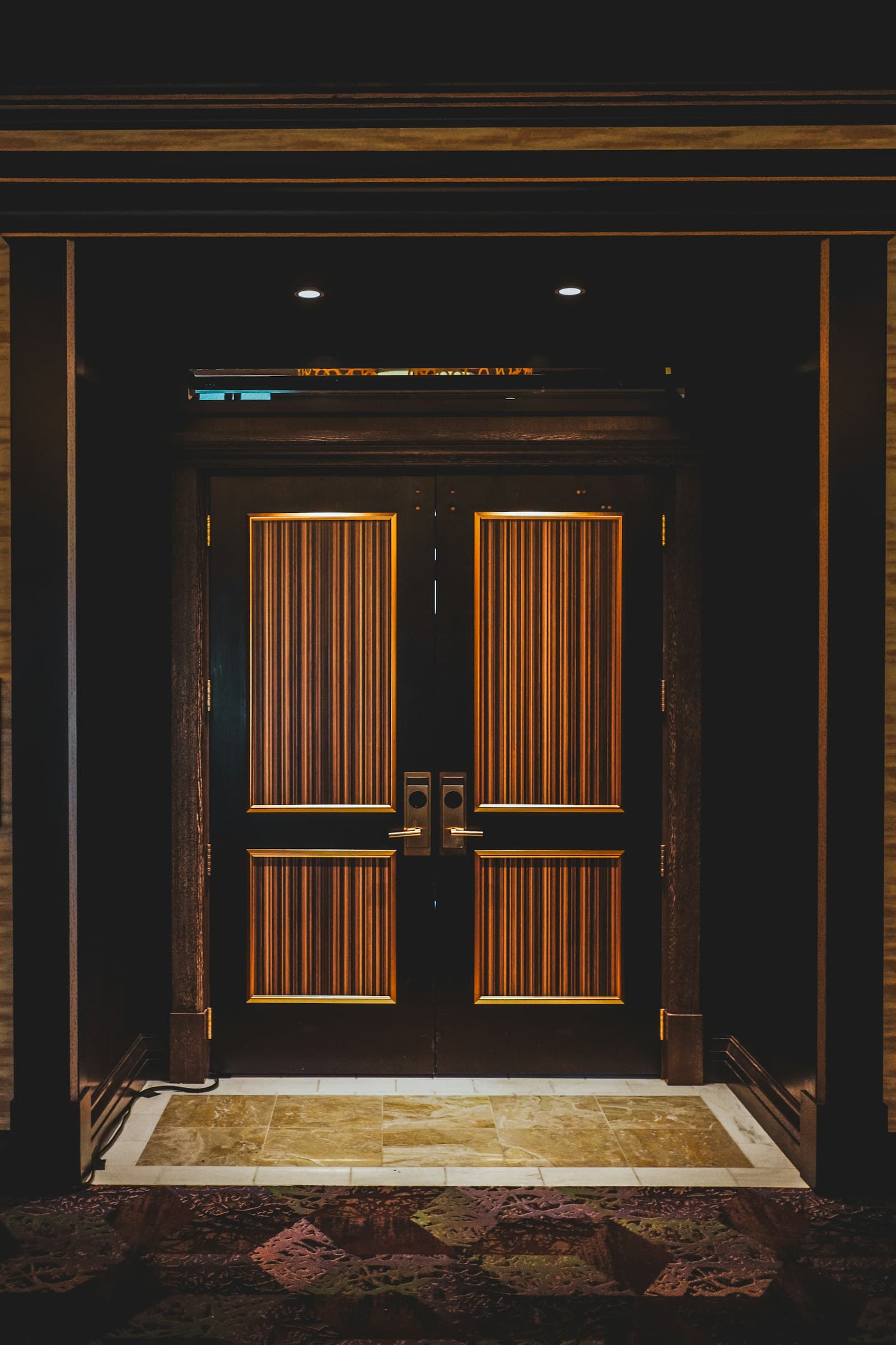 Puerta alta de doble hoja en madera oscura con incrustaciones de bronce, enmarcada por un portal panelado sobre un umbral de piedra