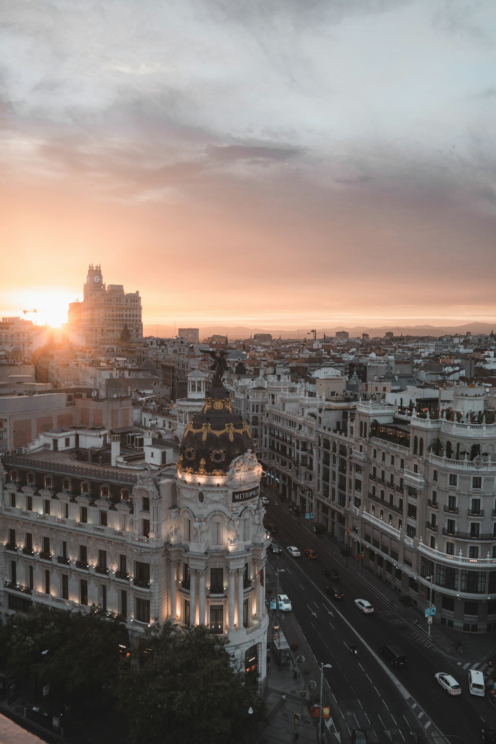 El centro de Madrid al anochecer, visto desde lo alto: un edificio en esquina coronado por una cúpula y una larga avenida bajo un cielo rosa pálido