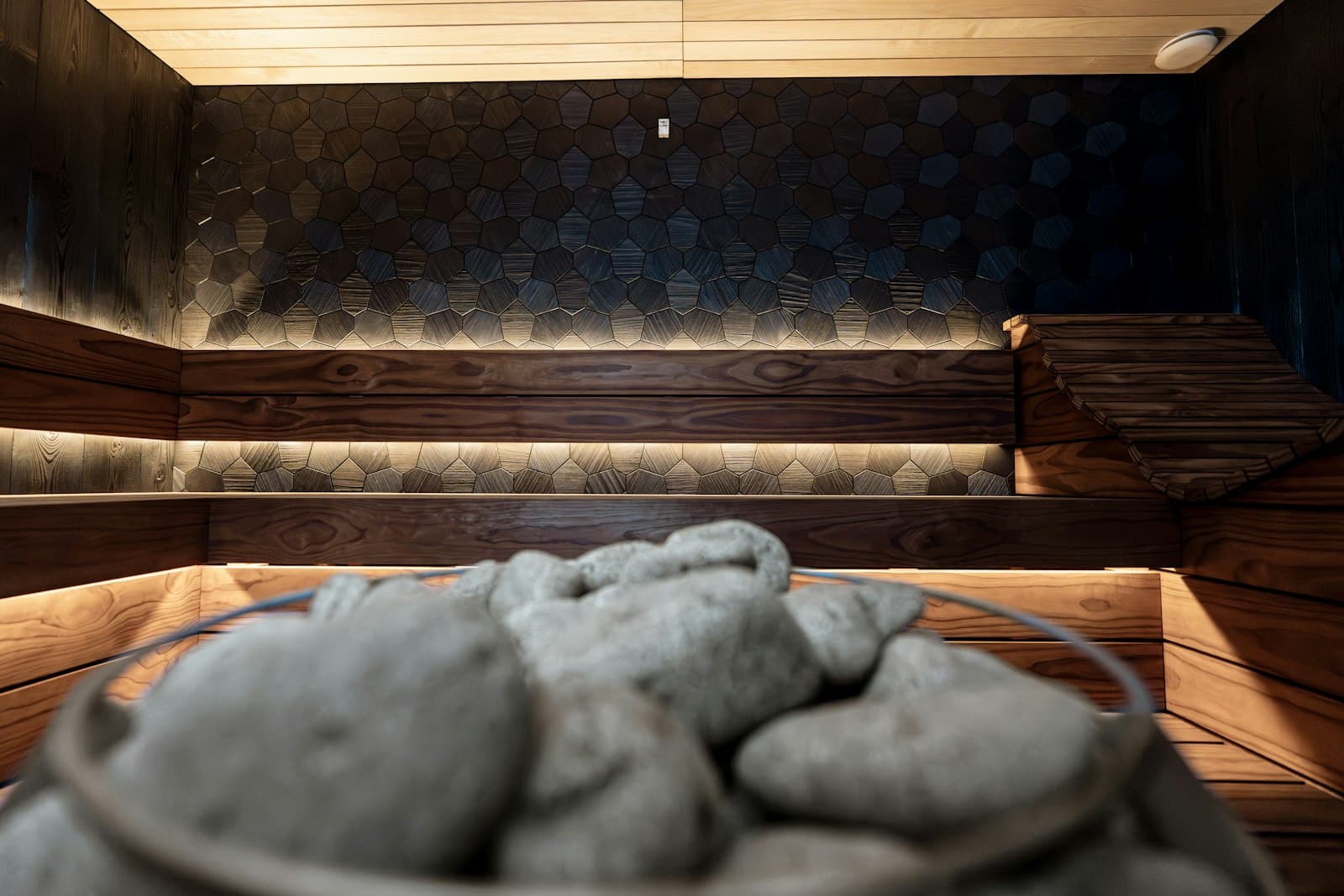 Piedras del calefactor en primer plano de una sauna de madera oscura, con una luz ámbar tenue bajo los bancos