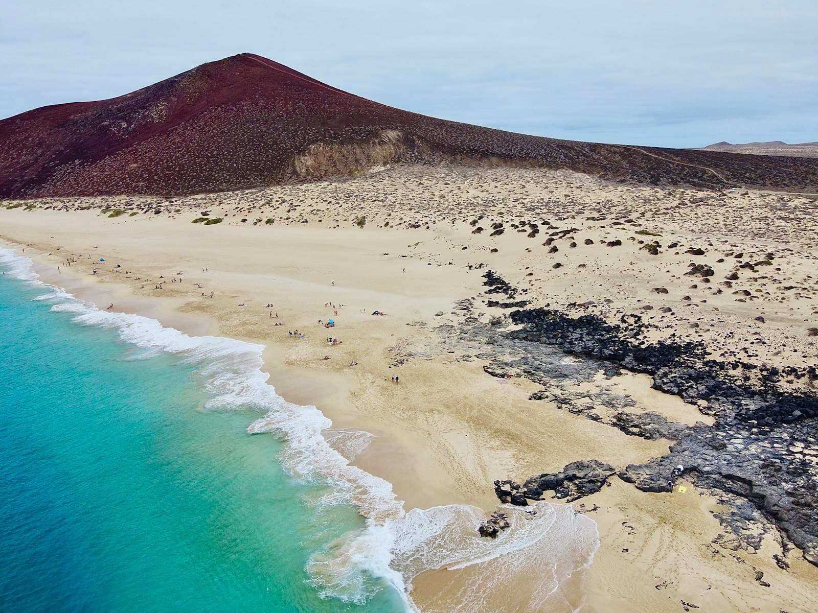 La bahía desde el aire: agua turquesa rompiendo sobre arena clara, al pie de una colina volcánica rojiza