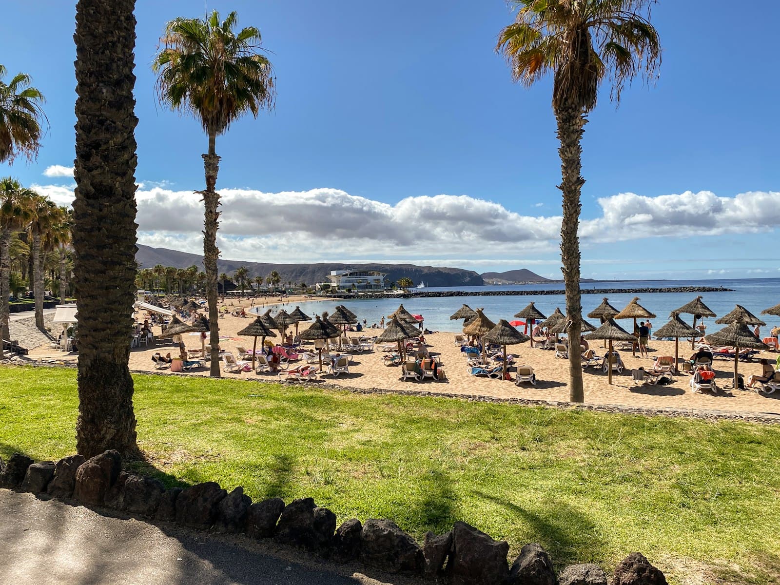 Playa Solmara vista desde el césped del jardín, con palmeras y sombrillas de paja sobre arena dorada