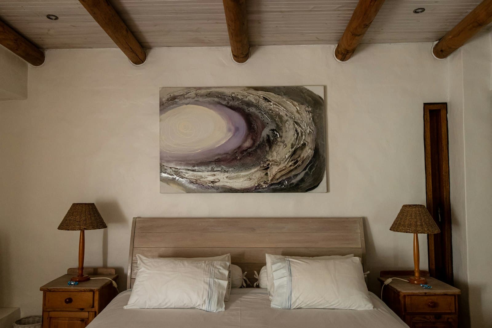 Cama de matrimonio con ropa blanca bajo vigas redondas de castaño, con lámparas de mimbre sobre mesillas de madera