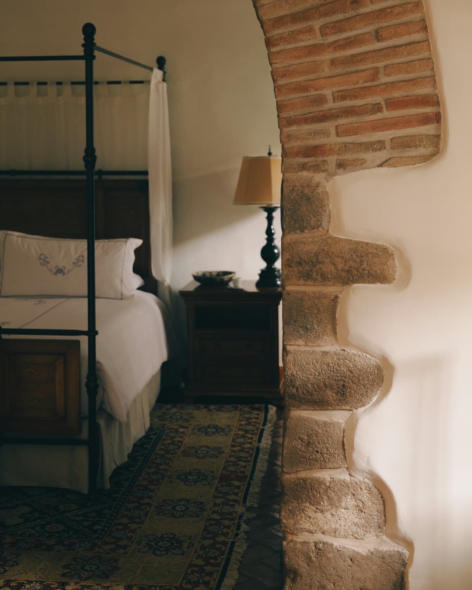 Cama de hierro con dosel y ropa bordada, vista a través de un arco de piedra y ladrillo